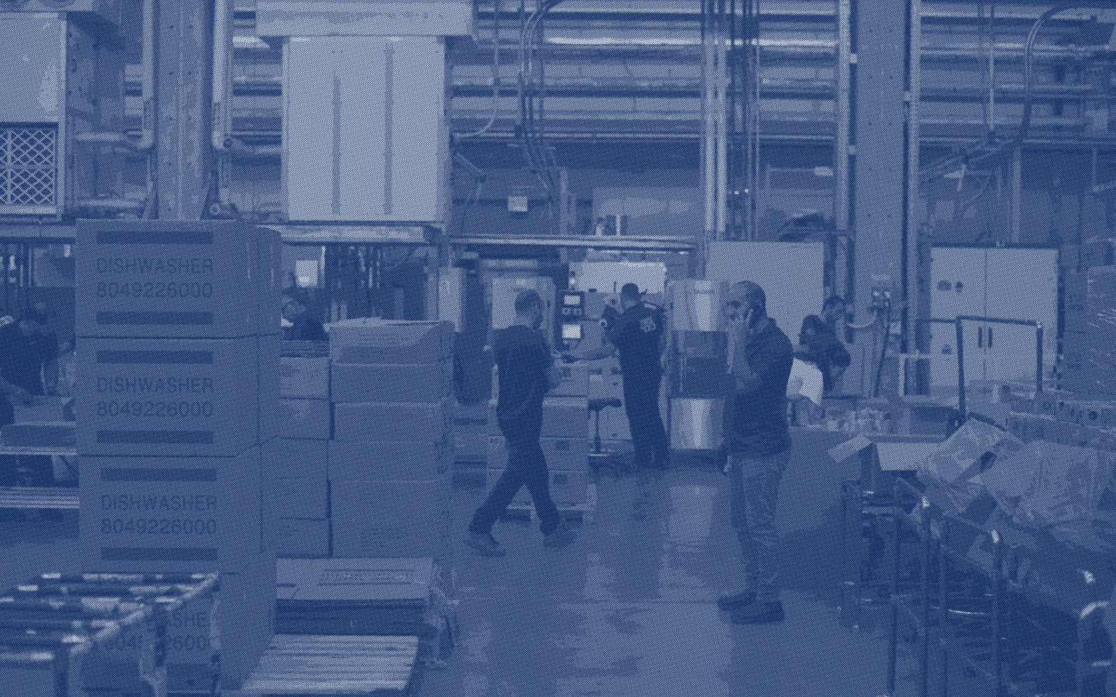 People crossing a plant floor between stacked pallets and machines