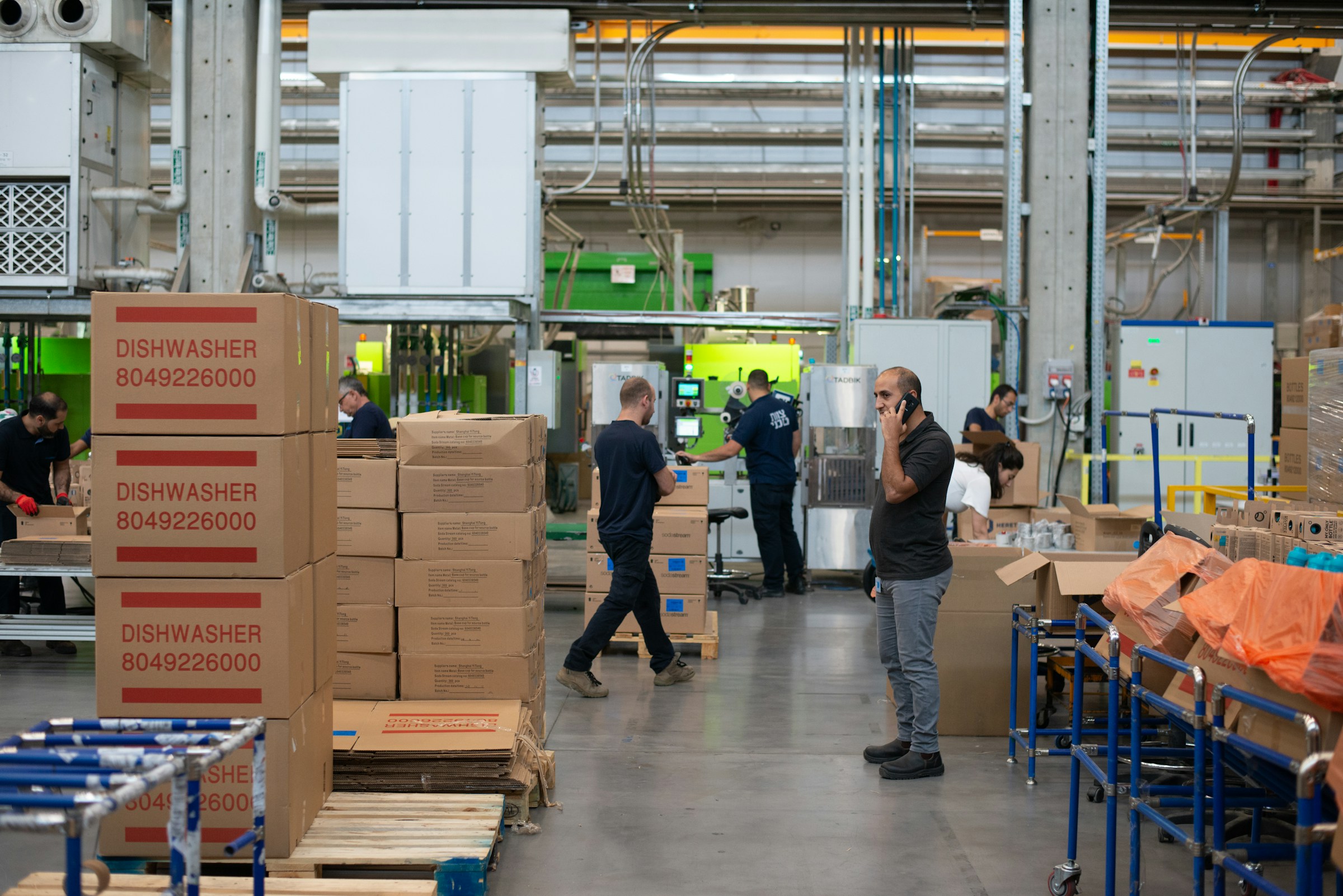 A production hall with cardboard boxes stacked on pallets, roll cages and people working at packing machines
